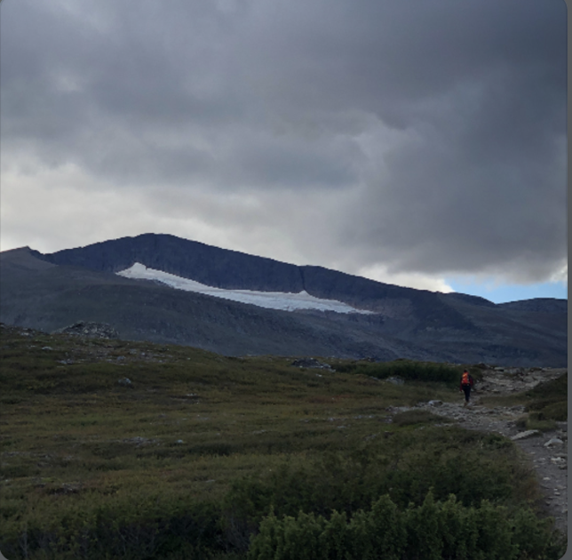 A person hiking on a rocky trail in a mountainous landscape, with a glacier visible on the mountain in the background under a cloudy sky.